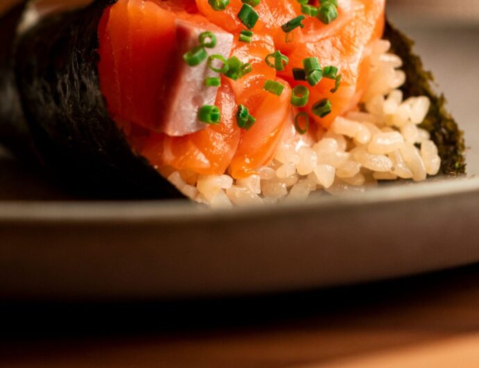 Close-up of a fresh salmon hand roll sushi garnished with chives on a ceramic plate with chopsticks.